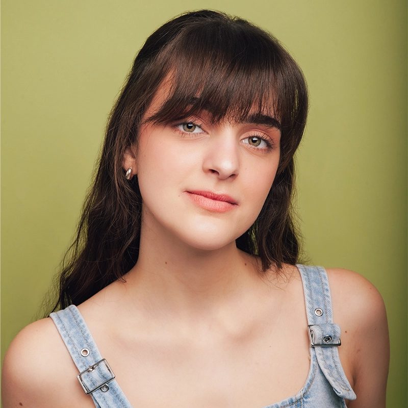 A young woman with long brown hair and bangs wears light makeup and denim overalls. She looks slightly to the side with a gentle, neutral expression. The soft lighting highlights her features against a plain pale green background—classic Alex Portaro style.