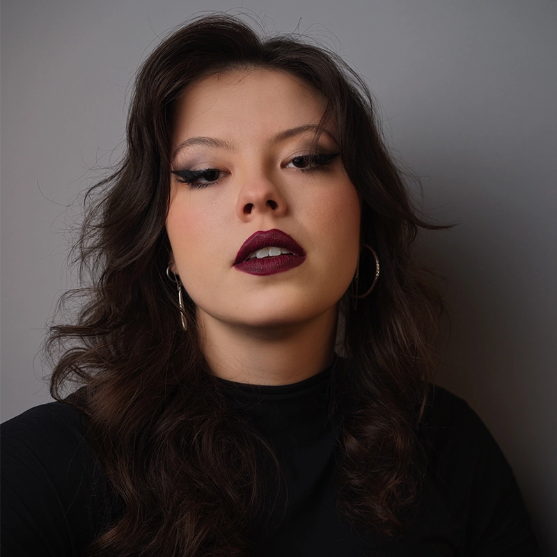 Anna Barrett, a young woman with wavy dark hair, bold dark lipstick, dramatic winged eyeliner, and hoop earrings, poses confidently in a black top against a plain gray background.