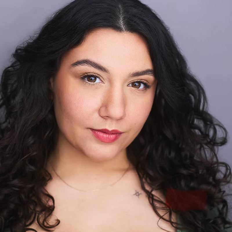 Diana Taylor, a woman with long, dark curly hair, poses against a plain gray background. She has light skin, brown eyes, neatly shaped eyebrows, natural makeup with pink lipstick, a small star tattoo near her collarbone, and a thin chain necklace.