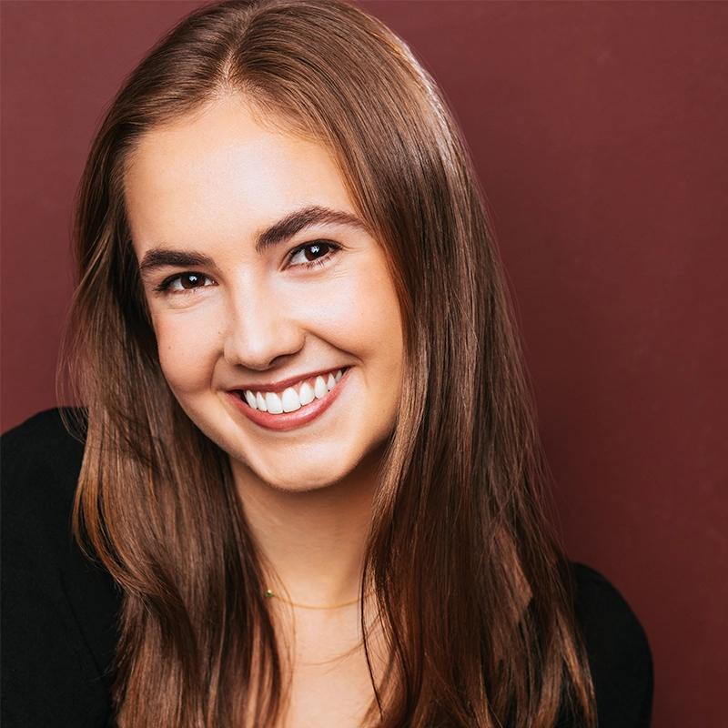 Anna Barrett, a young woman with long, straight brown hair, smiles warmly. She has light skin, brown eyes, and wears a black top. Set against a solid reddish-brown background, she faces the camera with a friendly, approachable vibe.
