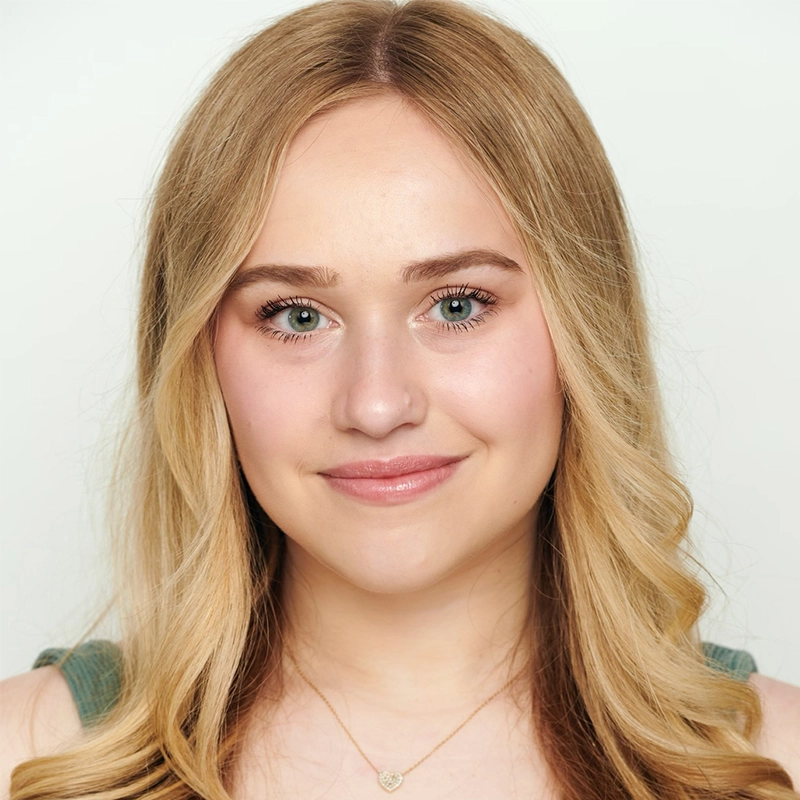 A young woman with long, wavy blonde hair smiles softly. She has blue eyes, light skin, and natural makeup. She wears a green top and a gold necklace with a round pendant, positioned against a plain light background.
