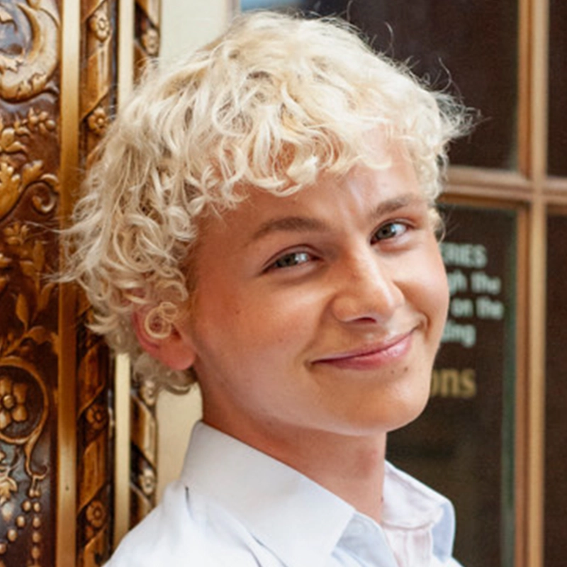 A young person with light skin and short, curly platinum-blonde hair smiles warmly. They wear a light blue collared shirt and stand near a decorative, ornately carved wooden panel with golden details. The background includes a blurred glass window.