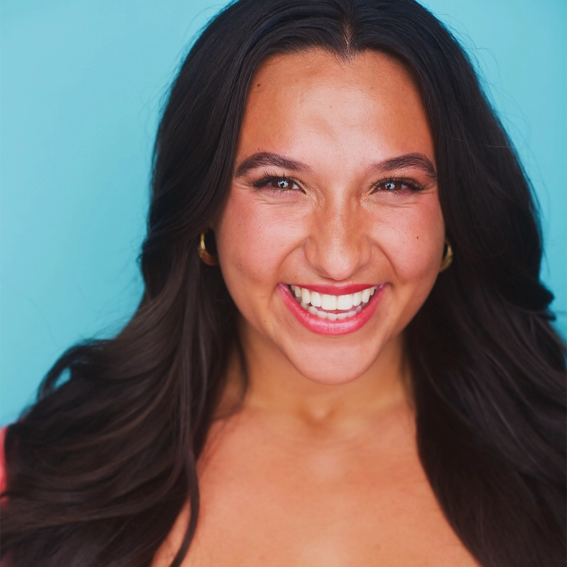 Marianna Ban, a woman with long dark hair, gold hoop earrings, and a pink top, smiles brightly at the camera. She has clear skin, natural makeup, and is posed against a solid light blue background.