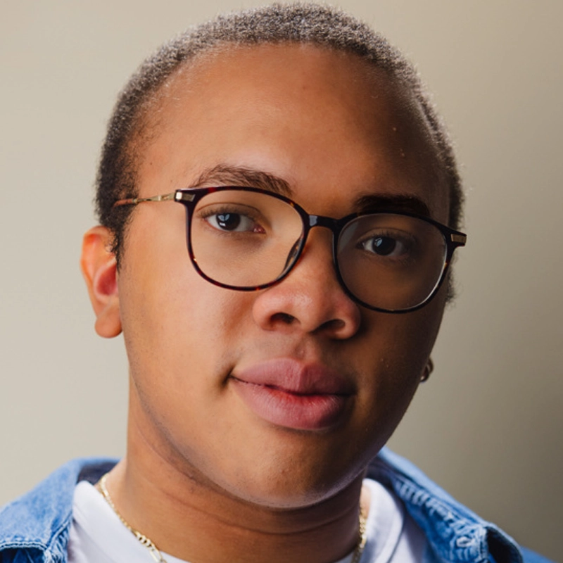 A person with short hair, round eyeglasses, and brown skin looks at the camera. They wear a blue denim shirt over a white top and a gold necklace. The background is plain and softly lit, highlighting their face—an image reminiscent of Bryson Battle’s signature style.