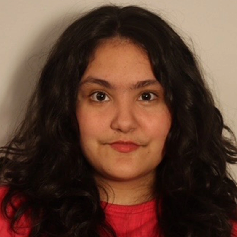 A person with long, wavy dark hair and fair skin is facing the camera. They are wearing a red top and have a neutral expression. The background is plain and light-colored.