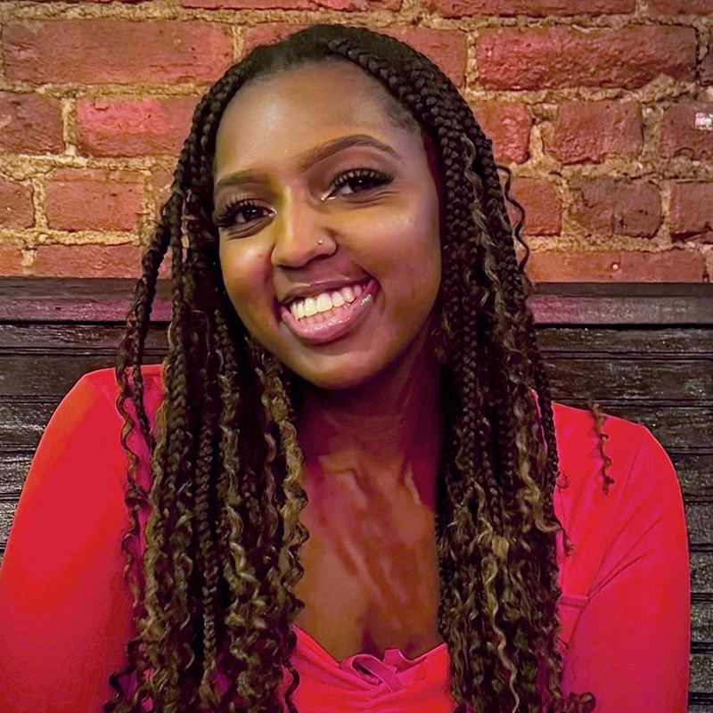 Carrington Symone, a woman with long, braided hair, smiles warmly at the camera. She is wearing a red top and sitting indoors against a rustic background of exposed brick and dark wooden panels in cozy, warm lighting.