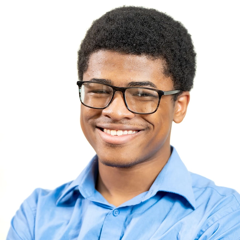 A young man with short curly hair and glasses smiles warmly. Wearing a light blue collared shirt, he stands against a plain white background, giving off a friendly and approachable Carrington Symone vibe.