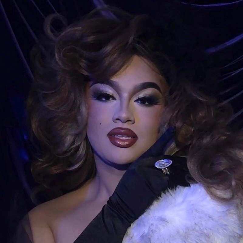 A glamorous person with voluminous, styled brown hair poses against a dark, silky backdrop. Channeling Diana Taylor, they wear dramatic makeup, dark lipstick, a jeweled ring atop a black glove, and a white fur stole—pure vintage Hollywood elegance.