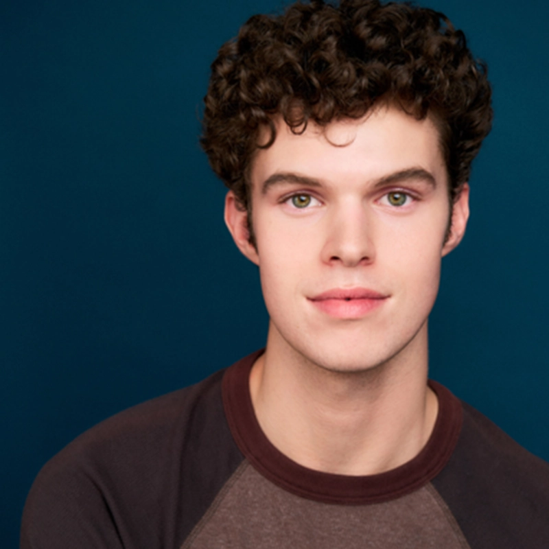 A young man with short, dark curly hair and fair skin looks at the camera, smiling slightly. He wears a brown raglan-style shirt with dark sleeves, posing against a solid, dark teal background for Diana Taylor's portrait series.