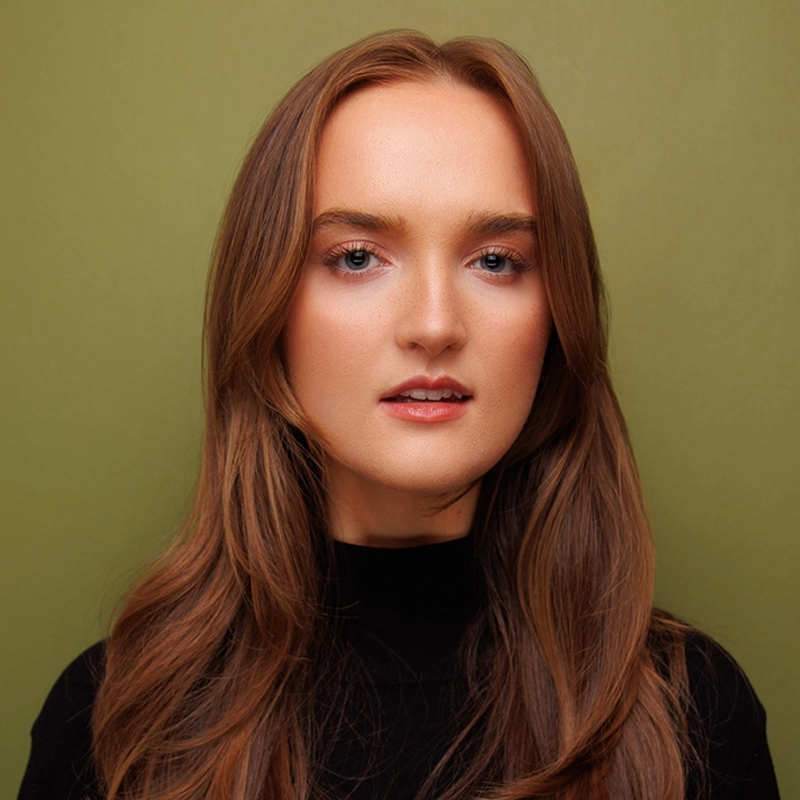 Carrington Symone, a young woman with long, light brown hair, poses in front of an olive green background. She has fair skin, subtle makeup, and wears a black top with a high neckline as she makes direct eye contact with the camera.