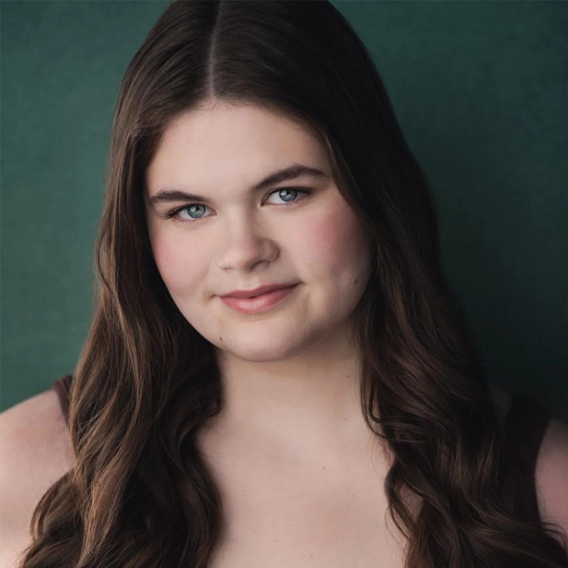 Carrington Symone, with long, wavy brown hair and fair skin, smiles softly at the camera. Her blue eyes and natural makeup stand out against the solid dark green background as she wears a sleeveless dark top.