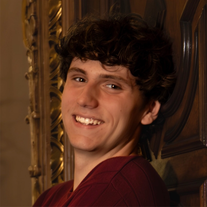 Tyler Landusky, a young man with wavy brown hair, smiles while leaning against an ornately carved wooden door. He wears a maroon shirt and looks directly at the camera as golden accents on the softly lit frame catch the light.