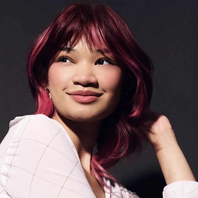 A person with shoulder-length, wavy magenta hair and curtain bangs smiles softly, looking to the side. Reminiscent of singer Angelica Hale from America's Got Talent, they wear a white, grid-patterned shirt and pose with one hand behind their head.