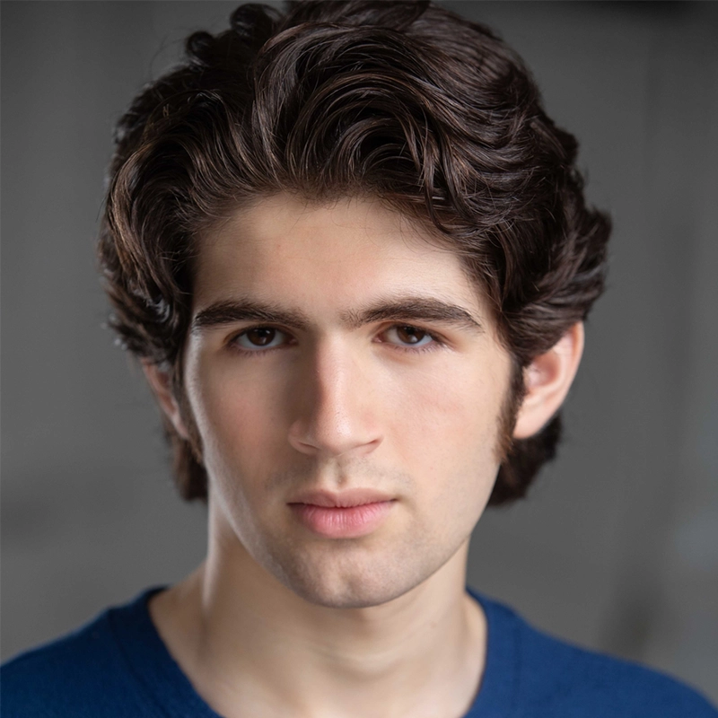 Carrington Symone, a young person with wavy dark brown hair and fair skin, looks directly at the camera with a neutral expression. Wearing a blue shirt against a blurred gray background, their face is the main focus.