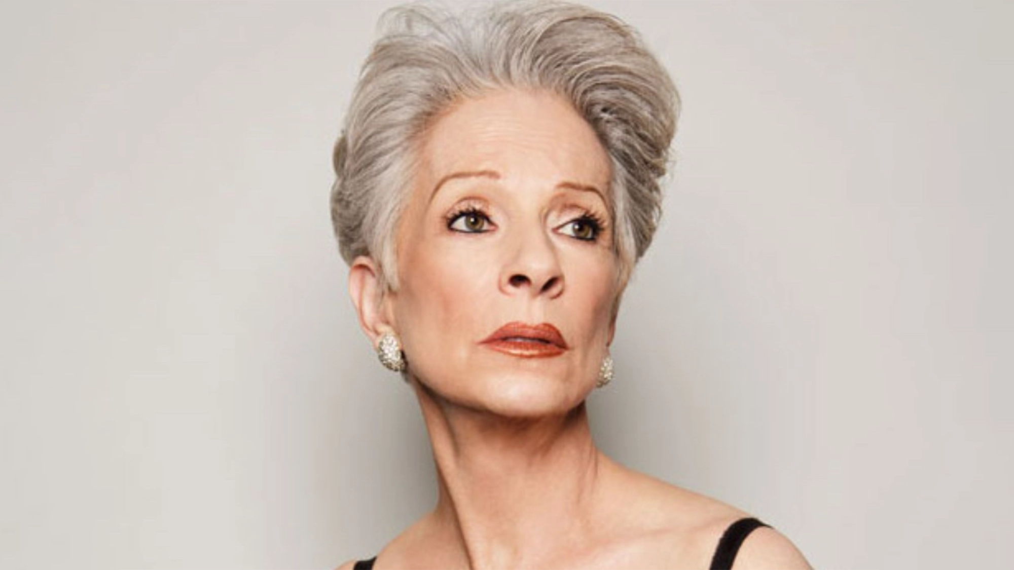 An elegant older woman, reminiscent of Jennifer Bassey, with short, styled gray hair gazes upward. She wears a sleeveless black top, pearl earrings, and subtle makeup. The plain, light background highlights her poised and confident expression.
