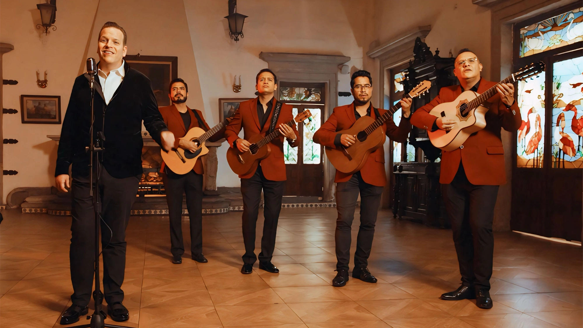 A five-member band performs indoors; four men in red blazers play guitars, while Jonathan Lolley, in a black jacket, sings into a microphone. The warmly lit room features stained glass windows and wood furnishings—perfect for a Hispanic Heritage Month celebration.