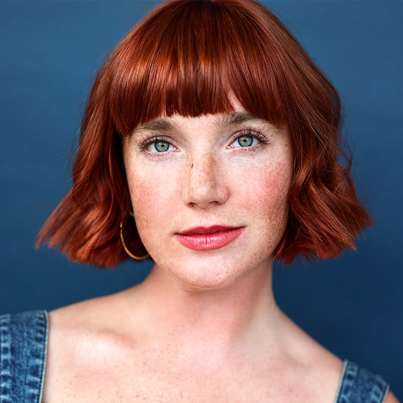 A woman with fair skin, short red hair with bangs, green eyes, and freckles looks at the camera. She wears natural makeup, gold hoop earrings, and a blue denim top. The background is a solid dark blue.