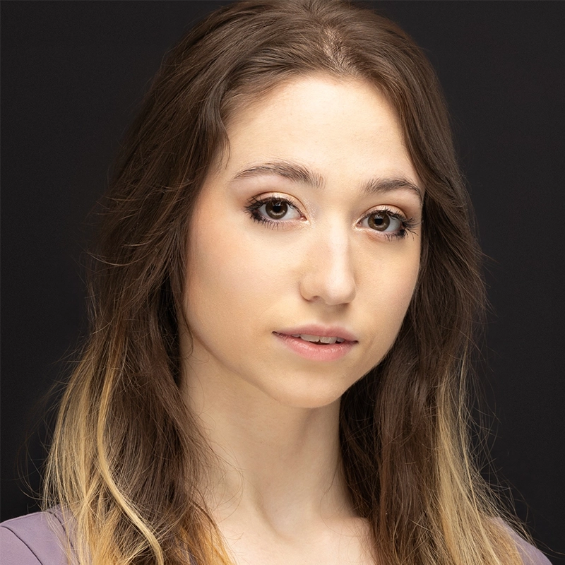 Carrington Symone, a young woman with long, wavy brown hair and subtle blonde highlights, poses against a dark background. She has fair skin, light brown eyes, and natural makeup, gazing calmly at the camera with a soft, neutral expression.