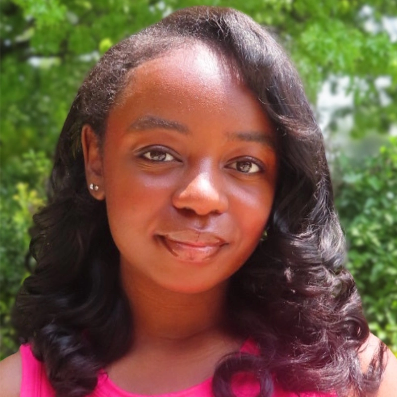A young Black woman with long, shiny, curled black hair poses outdoors, smiling softly. She wears a pink sleeveless top and small stud earrings. Green, leafy trees and bushes form a blurred natural background.