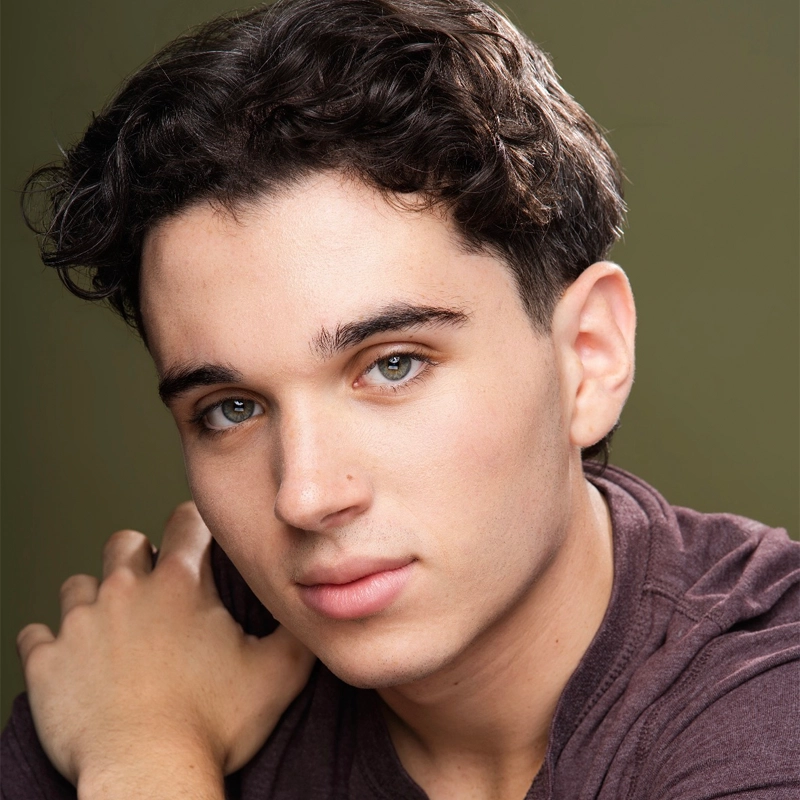 A young man with wavy dark hair and light eyes gazes softly at the camera. He wears a maroon shirt and rests one arm across his chest, with a neutral olive-green background behind him. The lighting highlights Tyler Landusky’s clear skin and facial features.