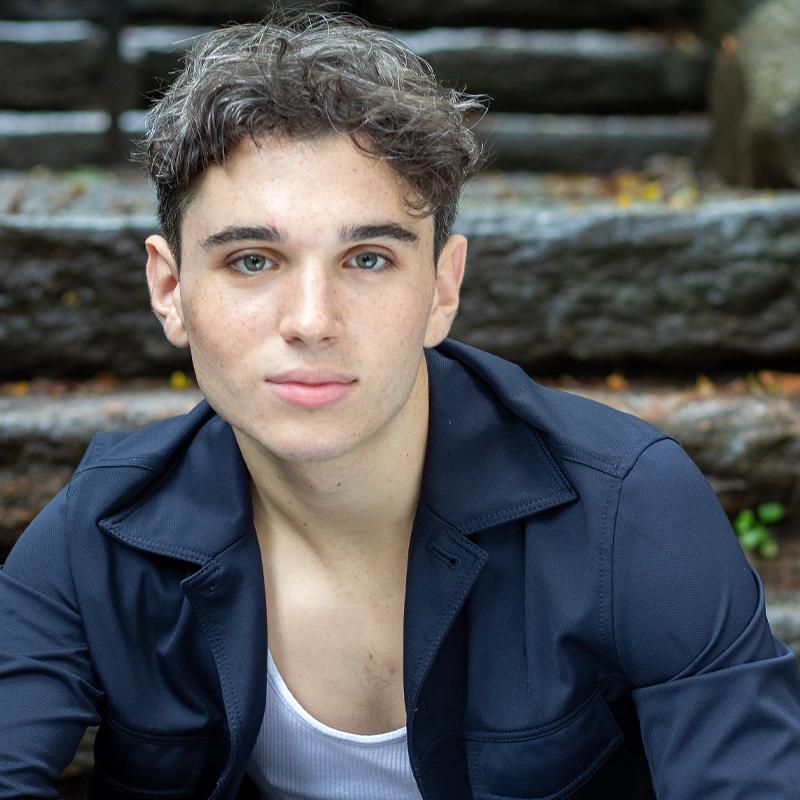 A young person with short, wavy brown hair and light eyes sits on outdoor stone steps. Tyler Landusky wears a navy jacket over a white shirt and looks directly at the camera with a neutral expression; hints of greenery blur in the background.