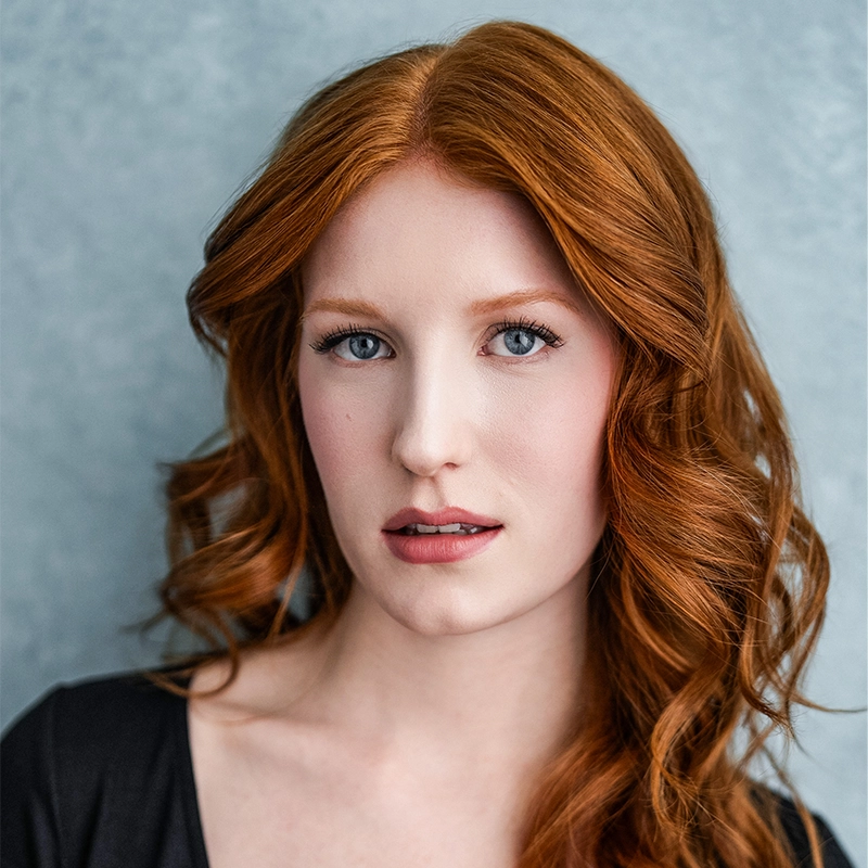 A headshot of a woman with vibrant, wavy copper-red hair parted in the center, framed against a textured gray background. She has piercing blue eyes, fair skin, and a neutral expression, wearing a simple black top. Her gaze is directed straight at the camera.