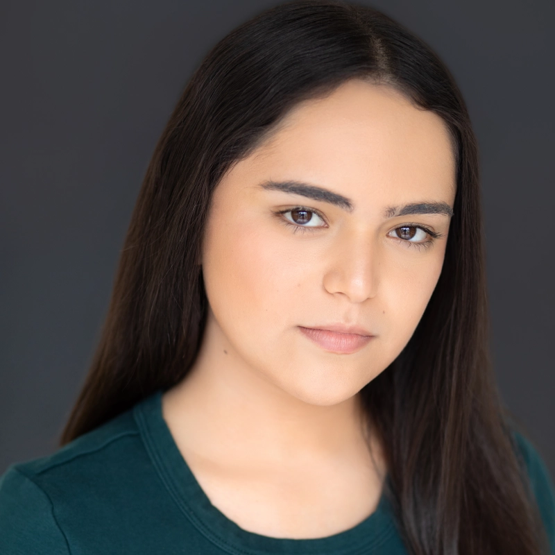 A young woman with long, straight dark brown hair and fair skin looks at the camera with a neutral expression. She wears a dark green top and stands against a plain, dark background. Her makeup is minimal and natural.