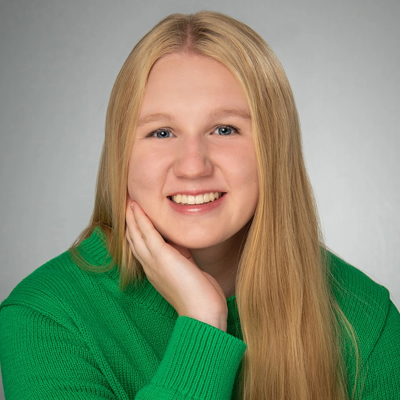 Carrington Symone, a young woman with long straight blonde hair, smiles warmly, resting her chin on her hand. She wears a bright green sweater and sits before a plain gray background, her expression friendly and approachable.