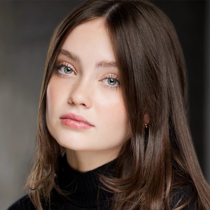 A young woman with fair skin, long straight brown hair, and light green eyes looks slightly to the side. She wears minimal makeup and a black turtleneck sweater, with a neutral expression, against a softly blurred dark background.