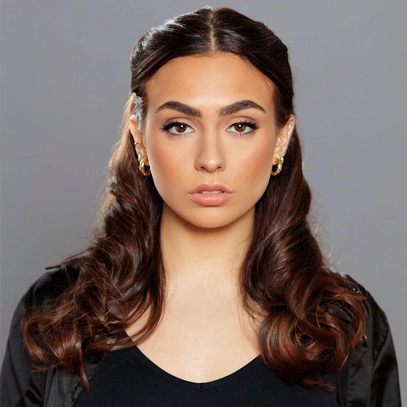 A woman with long, dark, wavy hair and bold eyebrows stares directly at the camera against a gray background. She wears gold hoop earrings, a black outfit, and a black jacket. Her makeup is natural with subtle highlighting and a nude lip color.