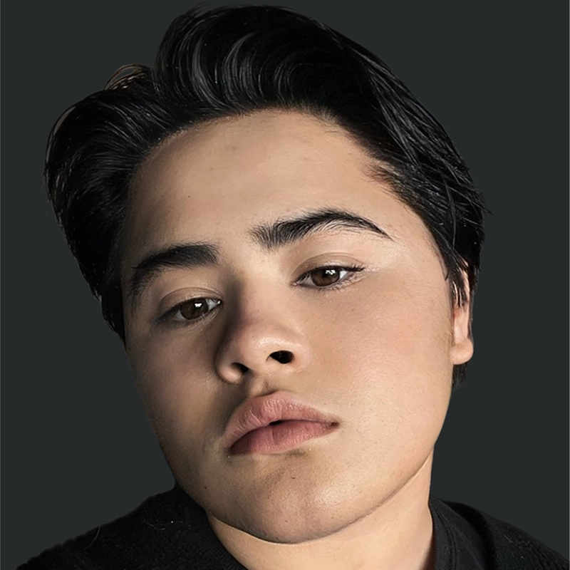 A person with short, dark hair and smooth, light skin looks directly at the camera with a neutral expression. They have well-groomed eyebrows, full lips, and are photographed against a plain, dark background.