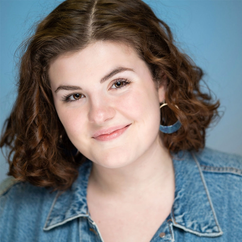 Young individual with curly brown hair, smiling gently. Wearing a blue denim jacket and circular blue earrings, Reagan White exudes charm. The background is a soft blue, complementing their casual and friendly expression perfectly.