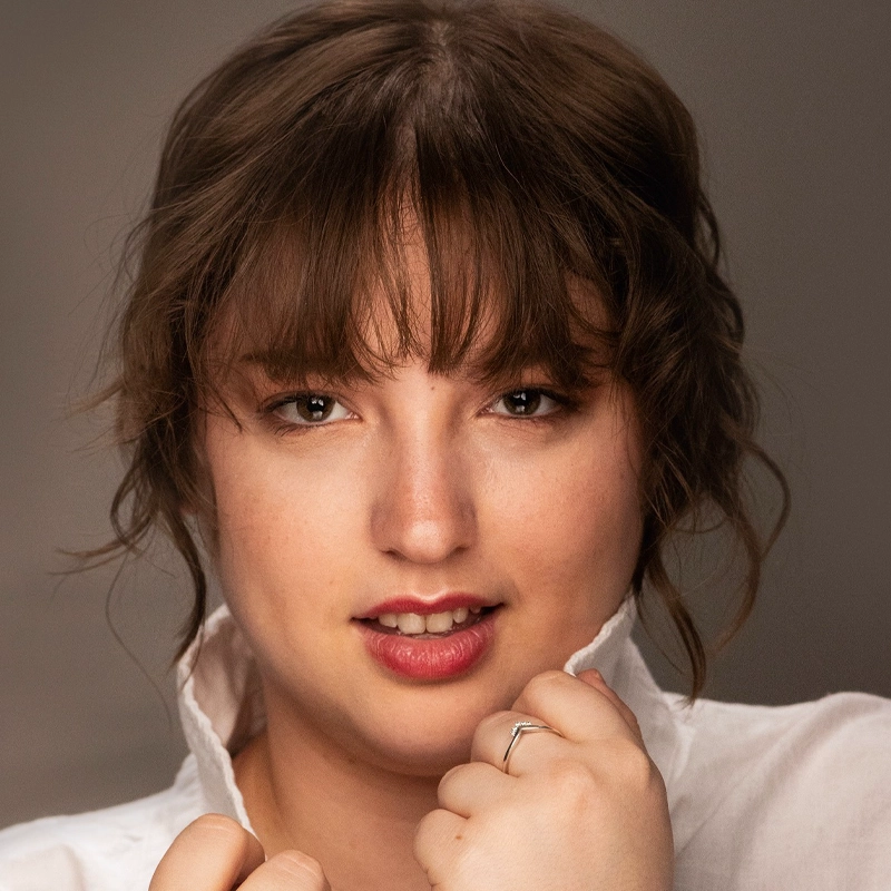 A young woman with wavy brown hair and bangs looks into the camera, wearing a white shirt with the collar turned up. She holds the collar with both hands, displaying a silver ring on her left hand. The background is neutral and softly lit.