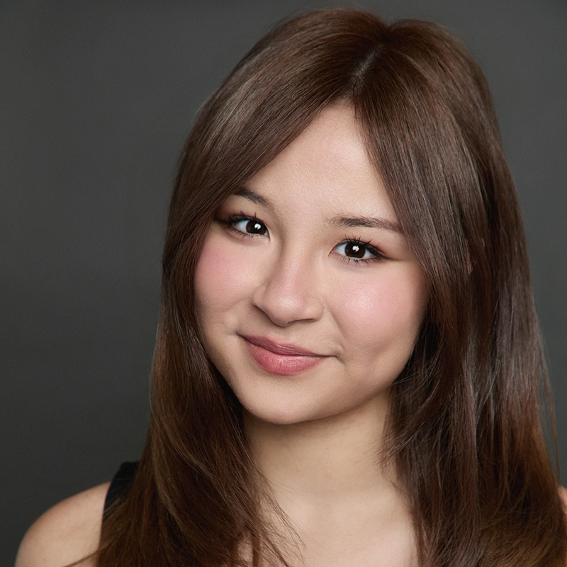 A young woman with straight, medium-length brown hair smiles softly. She has clear skin, natural makeup with pink blush, and is wearing a black top. The background is a plain dark gray, creating a professional studio portrait look.