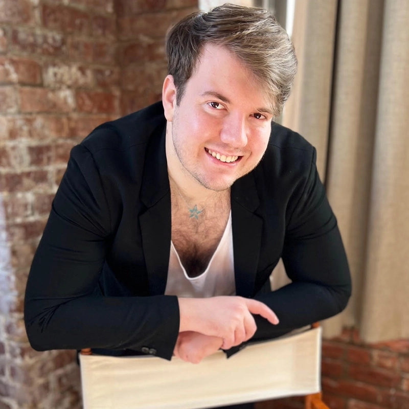 A young man with light brown hair and facial stubble leans forward on a white chair, smiling at the camera. He wears a black blazer over a white shirt, with a small chest tattoo visible. The background includes brick walls and beige curtains.