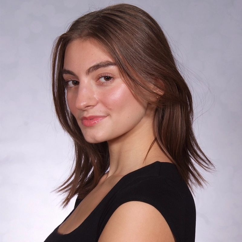 A young woman with light skin and straight, shoulder-length brown hair wears a black top. She faces slightly to the side, smiling softly. The softly blurred, light gray background gives a clean, studio portrait look by Tyler Landusky.