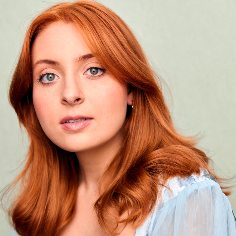 Natalie Alexa Taylor, a young woman with long, wavy red hair and fair skin, looks slightly past the camera. She has blue eyes, subtle makeup, and wears a sheer light blue top against a softly lit background for a calm, natural feel.