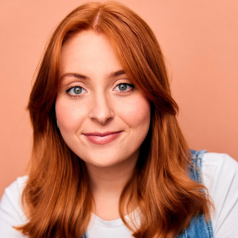 Natalie Alexa Taylor, a young woman with long, wavy red hair and light blue eyes, smiles softly. She wears a white shirt with a light blue vest against a solid peach background, posing in a friendly, relaxed manner.