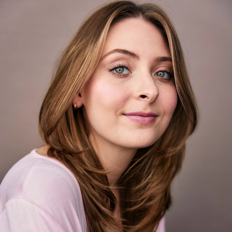 A young woman, Natalie Alexa Taylor, with light skin, blue eyes, and straight, light brown hair smiles gently at the camera. She wears a pale pink top and small hoop earrings. A soft, neutral background highlights her relaxed confidence.
