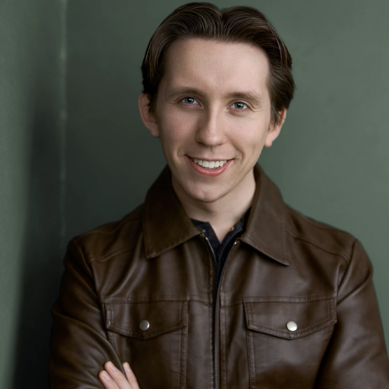 A smiling person, Bradley Viktor, with short brown hair wears a brown leather jacket over a dark shirt. They stand against a muted green wall with arms crossed, looking directly at the camera. The lighting is soft, highlighting their face and jacket.