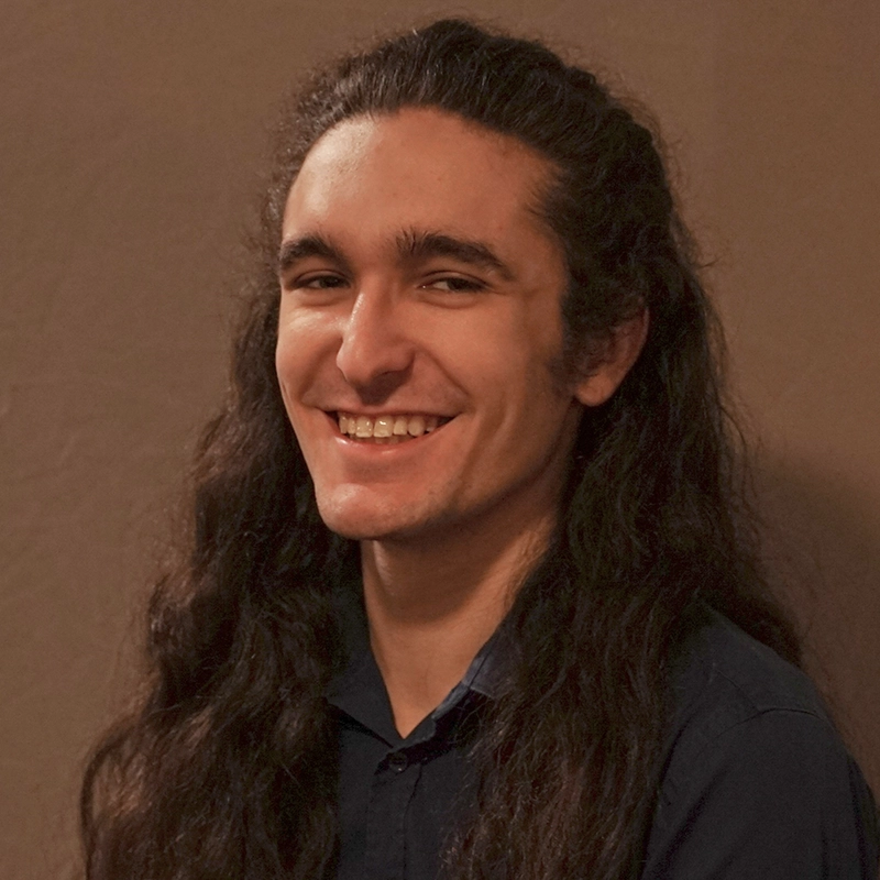 A young person with long, wavy dark hair and light skin smiles at the camera. Sawyer Whitted is wearing a dark collared shirt and is posed against a plain, brown background. The soft lighting highlights their cheerful expression.