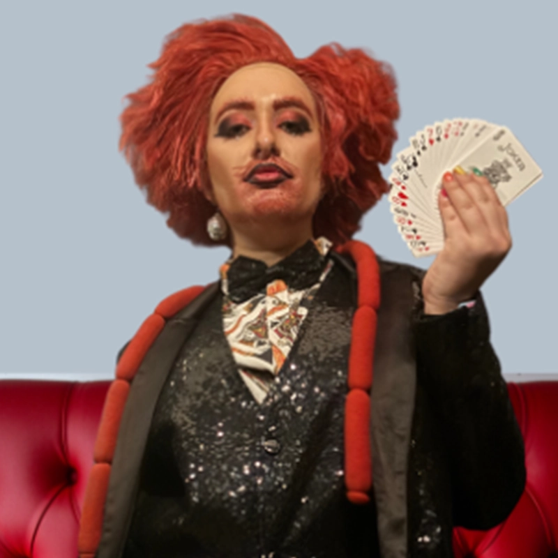 A person with red, styled hair and dramatic makeup wears a sparkling black suit, patterned bow tie, and large fake sausage necklace, sitting on a red tufted couch, holding a fan of playing cards in one hand and posing confidently.