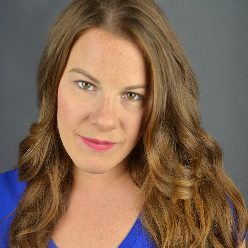 A woman with long, wavy light brown hair wearing a blue top looks into the camera with a slight smile. She has fair skin, light makeup, and stands against a plain gray background.