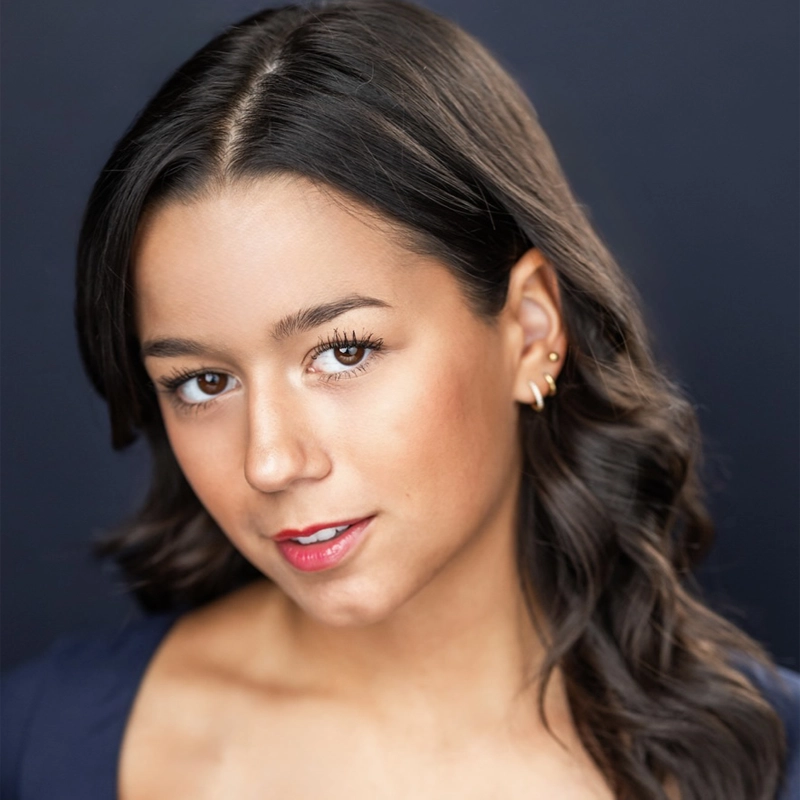 Makaila Anderson, a young woman with wavy dark brown hair and warm skin tone, gazes at the camera. She wears gold earrings, subtle makeup with red lipstick, and a dark top. A soft, dark gradient background emphasizes her confident expression.