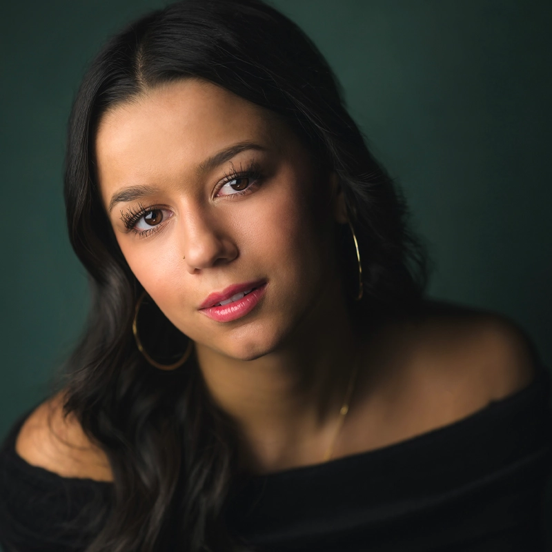 Makaila Anderson, a young woman with long dark hair, wears gold hoop earrings and a black off-the-shoulder top as she poses against a dark green background. She has a calm expression, soft makeup, and slightly glossy pink lips.