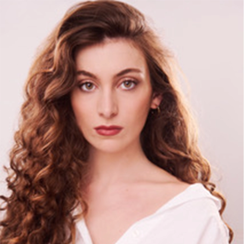Ava Longarzo, a young woman with long, wavy brown hair and fair skin, gazes directly at the camera with a neutral expression. She wears a white off-the-shoulder blouse and small hoop earrings against a soft, light background.
