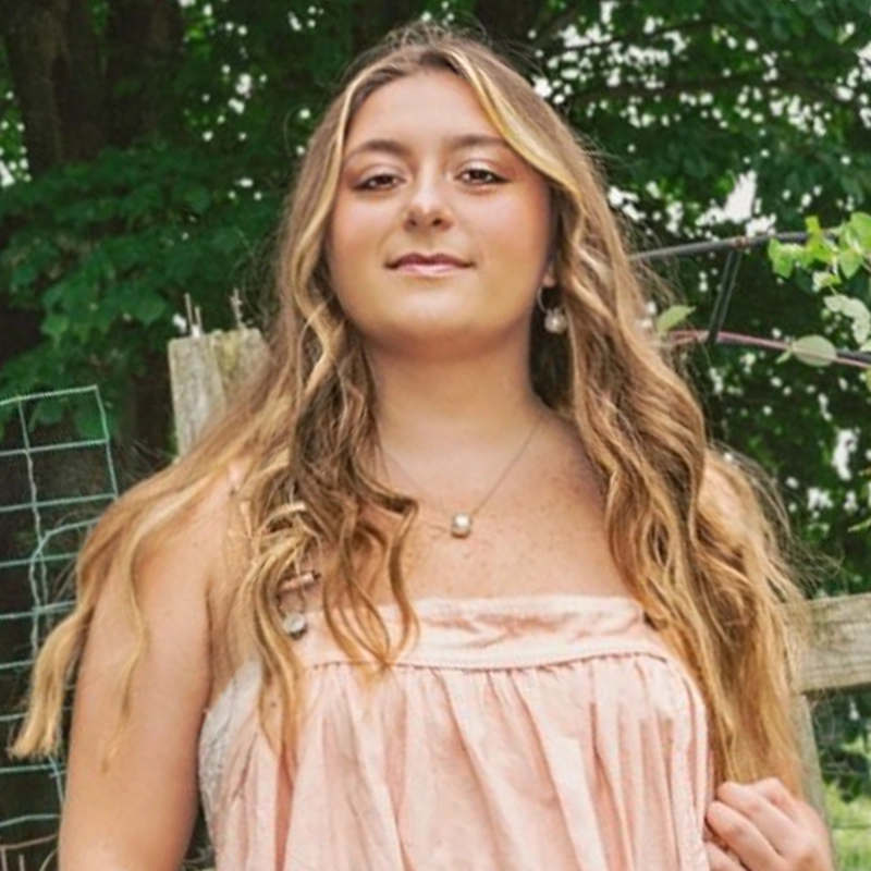A young woman with long wavy blonde hair wears a strapless pale pink dress and a pearl necklace, standing outdoors. Green trees, a wire fence, and wooden posts are in the background, giving a summery and rustic feel to the scene.