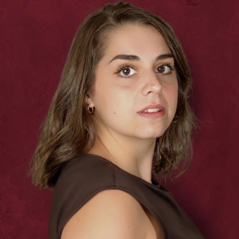 Young woman with shoulder-length brown hair, wearing a sleeveless dark top, stands in front of a maroon background. Olivia Brice faces slightly to the side, looking toward the camera with a neutral, soft expression. Lighting is even, highlighting her facial features.