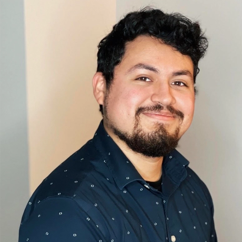 Christopher Quintana, a man with short, curly black hair and a beard, is smiling at the camera. He is wearing a dark navy shirt with a subtle dotted pattern and stands against a plain, light-colored background.