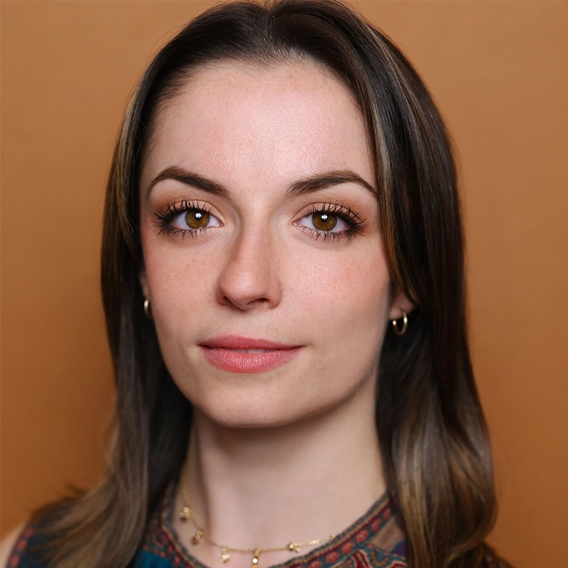 A young woman with fair skin and straight brown hair, parted in the middle, looks directly at the camera. Natalie O'Leary wears small hoop earrings, a delicate necklace, and a patterned top against a warm tan background.